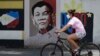 FILE - A woman wearing a protective mask rides her bicycle past an image of Philippine President Rodrigo Duterte in Manila, Philippines, on March 20, 2020. 